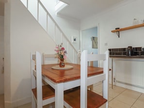 Un comedor con una mesa y sillas de madera, un jarrón con flores, una escalera y una zona de cocina con un estante.