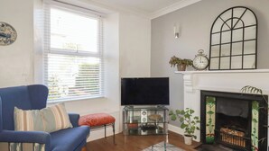 A living room with a blue sofa, a flat-screen TV, a fireplace, and a window with blinds. - Tin Lantern Cottage (Redruth)