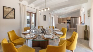 A modern dining room with a glass table, yellow chairs, and a kitchen in the background. - Son Barba Mallorca – Luxury, Privacy, Private Pool & Jacuzzi, AC, BBQ, More (Sa Pobla)