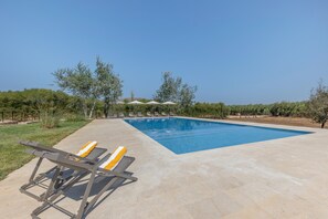 Una piscina rectangular con superficie de agua azul cristalina, rodeada por una plataforma de hormigón. Se observan sillones de relax con cojines amarillos y una toalla blanca a un lado.