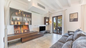 A modern living room with a large sectional sofa, a flat-screen TV, a glass display case with books, and a large window with a view of the outdoors. - Son Barba Mallorca – Luxury, Privacy, Private Pool & Jacuzzi, AC, BBQ, More (Sa Pobla)