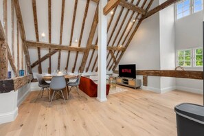 A loft-style room with exposed wooden beams, a dining area with chairs, a TV, and a window with a view. - Modern & Historic Duplex Flat Riverside (Surrey)