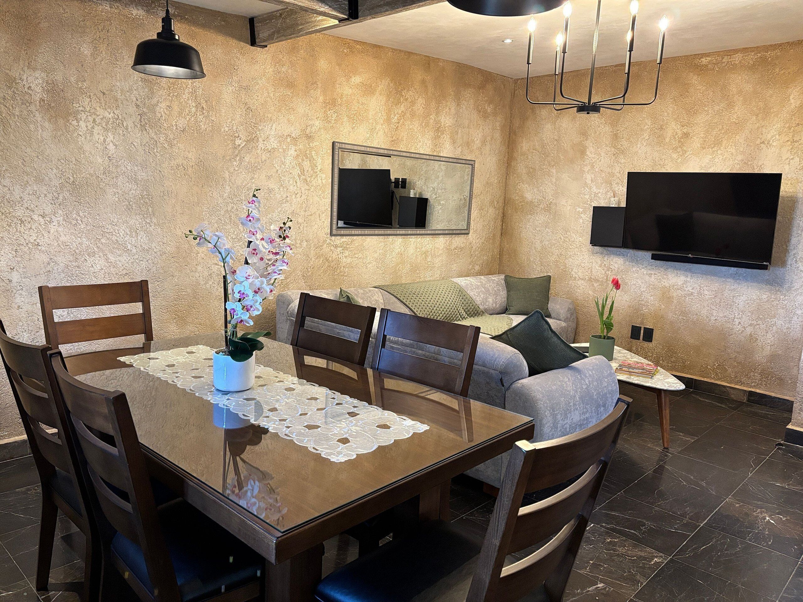 Un área de comedor con una mesa de madera, sillas, un jarrón de flores, un sofá, un televisor montado en la pared y una mesita auxiliar con una rosa.