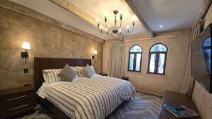 Habitación de hotel con una cama grande, cabecera de madera, dos lámparas de noche, un candelabro, un televisor y dos ventanas con cortinas.