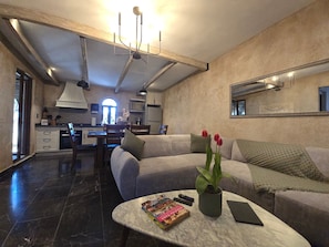 Una sala moderna con un sofá, una mesa de centro con libros y un control remoto, una planta en maceta y una cocina al fondo.