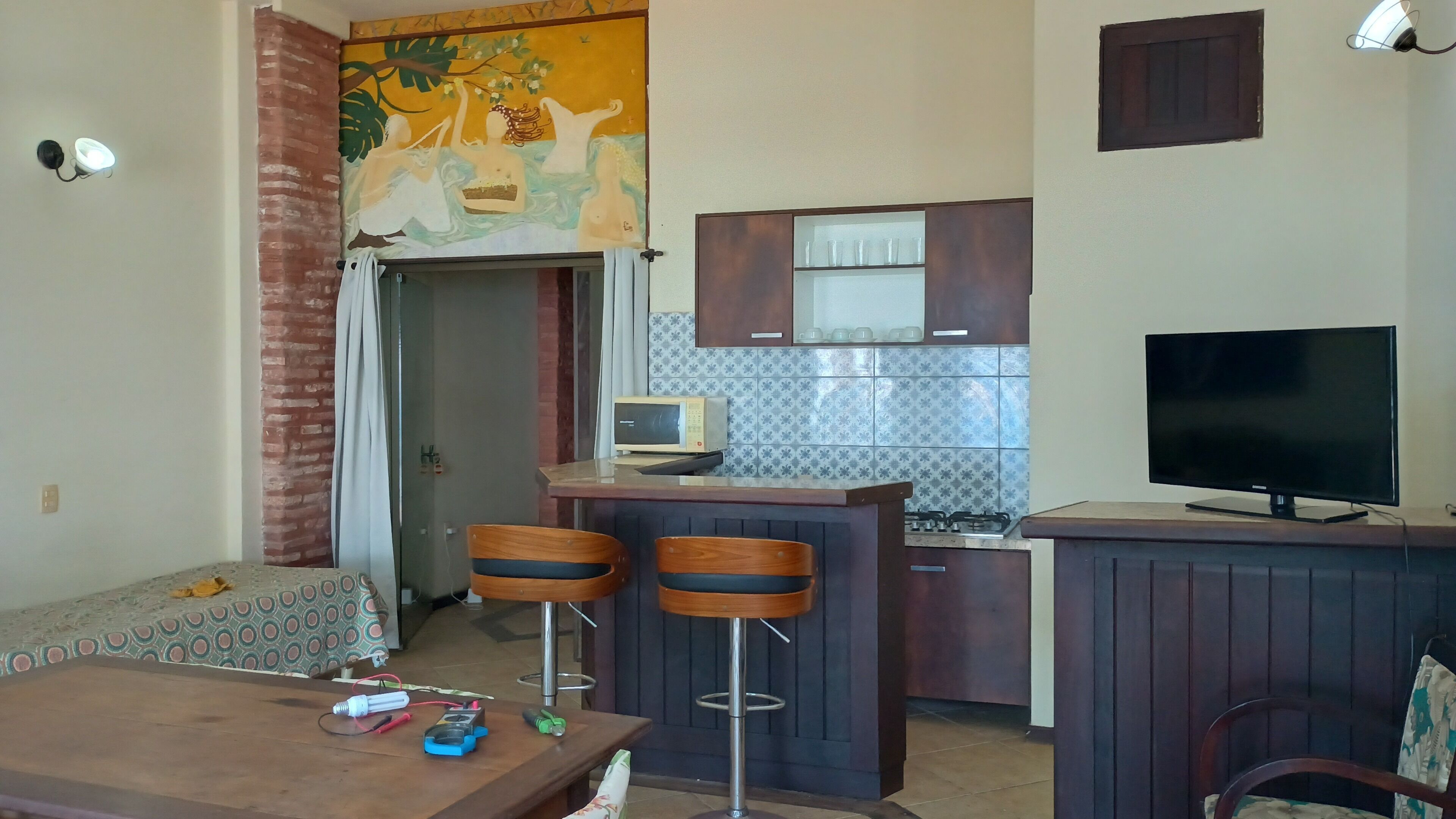 Un espacio habitable compacto con kitchenette, una zona de comedor con dos taburetes altos, un televisor y un mural en la pared.