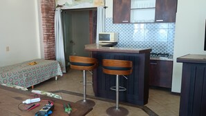 A compact kitchen with a microwave, a small sink, and a stove. There are two wooden bar stools and a bed with a patterned bedspread. - Direita S.Antonio-Plus Quarto e sala para 5 pessoas, vista baia Todos os Santos (Salvador)
