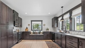 A modern kitchen with dark wood cabinets, a built-in bench with cushions, a large window, and a view of greenery outside. - Group Retreat Near Glacier | Lake Views (Somers)