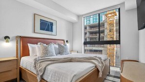 A modern bedroom with a large bed, a bedside table, a nightstand, a framed picture on the wall, and a window with a view of construction. - The Crosby Miami Worldcenter (Miami)