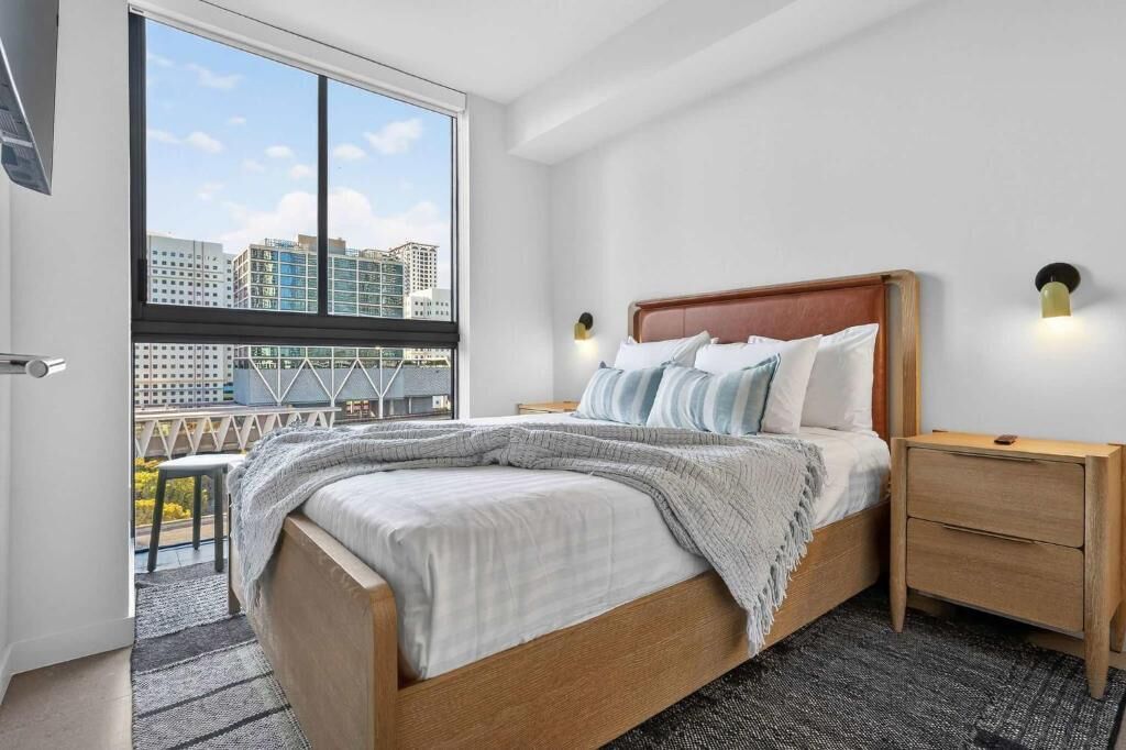 A bedroom with a large window, a bed with a wooden headboard, two bedside tables, and a view of buildings.