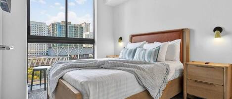 A bedroom with a large window, a bed with a wooden headboard, two bedside tables, and a view of buildings.