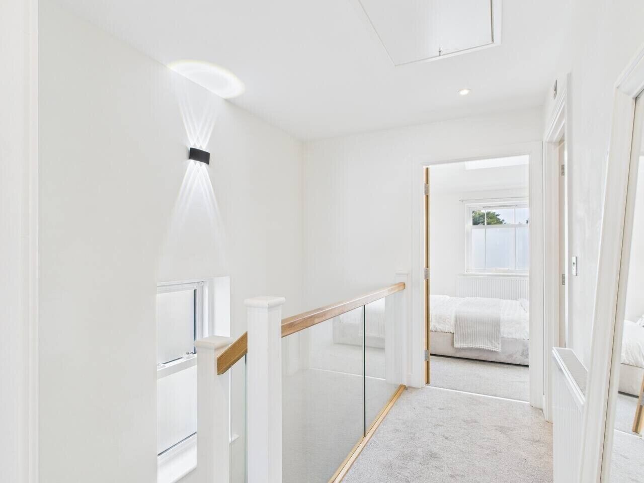 Un pasillo moderno y minimalista con barandilla de cristal, techo blanco y vista a un dormitorio con cama y ventana.