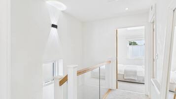 Un pasillo moderno y minimalista con barandilla de cristal, techo blanco y vista a un dormitorio con cama y ventana.