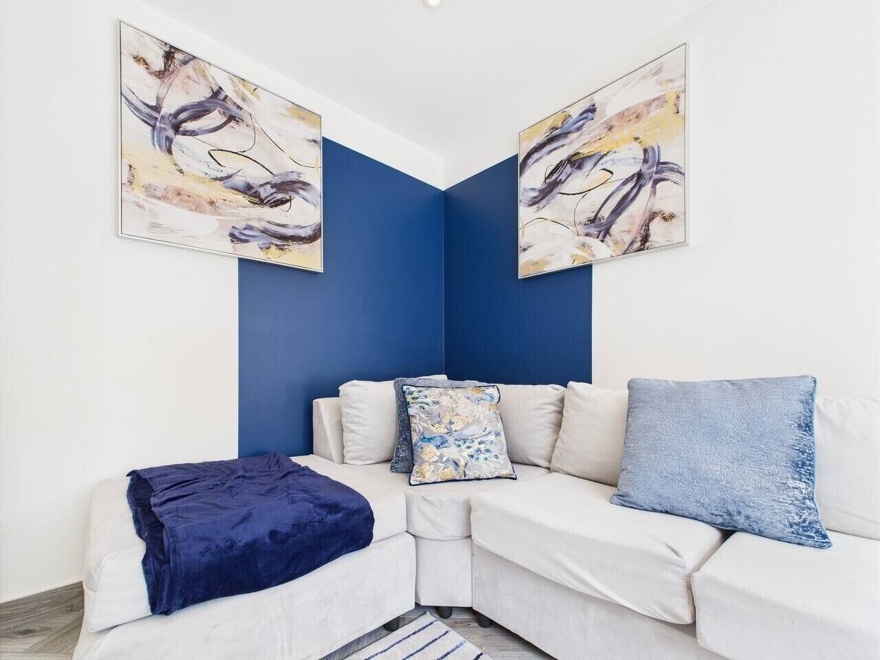 Een hoekwoonkamer met een witte bank, blauwe kussens, een blauwe plaid en abstracte kunst aan de muren.