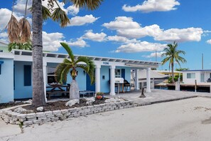 A coastal building with a blue facade, numbered 11296, featuring a covered outdoor seating area and a small garden with palm trees. - La Pirata: Hgtv-Remodeled 3BR/2BA Canal Home w/ Pool (Marathon)