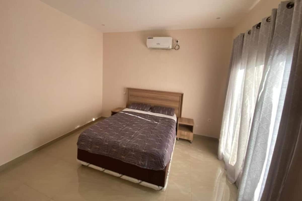 Un dormitorio con cama, mesita de noche, aire acondicionado de pared y ventana con cortinas.