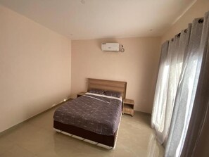 Un dormitorio con cama, mesita de noche, aire acondicionado de pared y ventana con cortinas.
