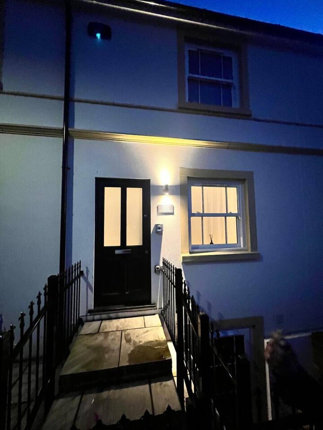 Una casa de dos pisos con una puerta principal negra, un ventanal con cortinas y una luz encima de la puerta.