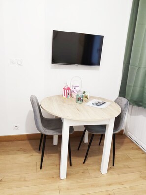 Ein runder Holztisch mit vier Stühlen, ein an der Wand befestigter Flachbildfernseher und ein grüner Vorhang auf der rechten Seite.
