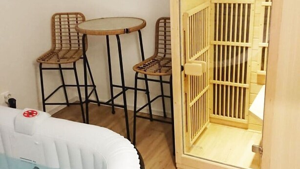 Ett rum med ett litet runt bord, två fåtöljer i rotting, en vit uppblåsbar säng och en trädörr med ett fönster med galler.