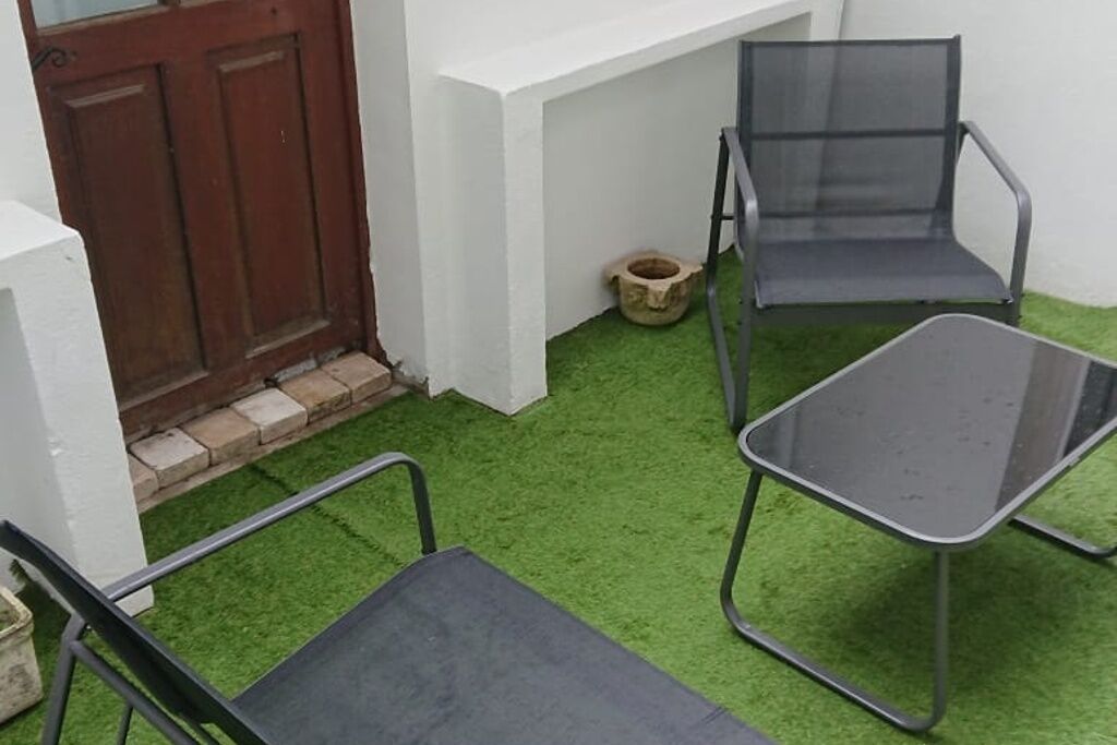 A small outdoor area with artificial green turf, two metal chairs, a metal table, a wooden door, and a white wall.