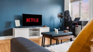 Una sala de estar con una televisión mostrando 'NETFLIX', un ventilador, una cinta de correr y un ventanal con vistas.