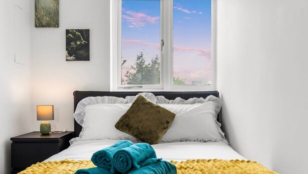 Una cama bien hecha con toallas enrolladas, una mesita de noche con lámpara y una ventana que ofrece vistas a árboles y un cielo rosado.