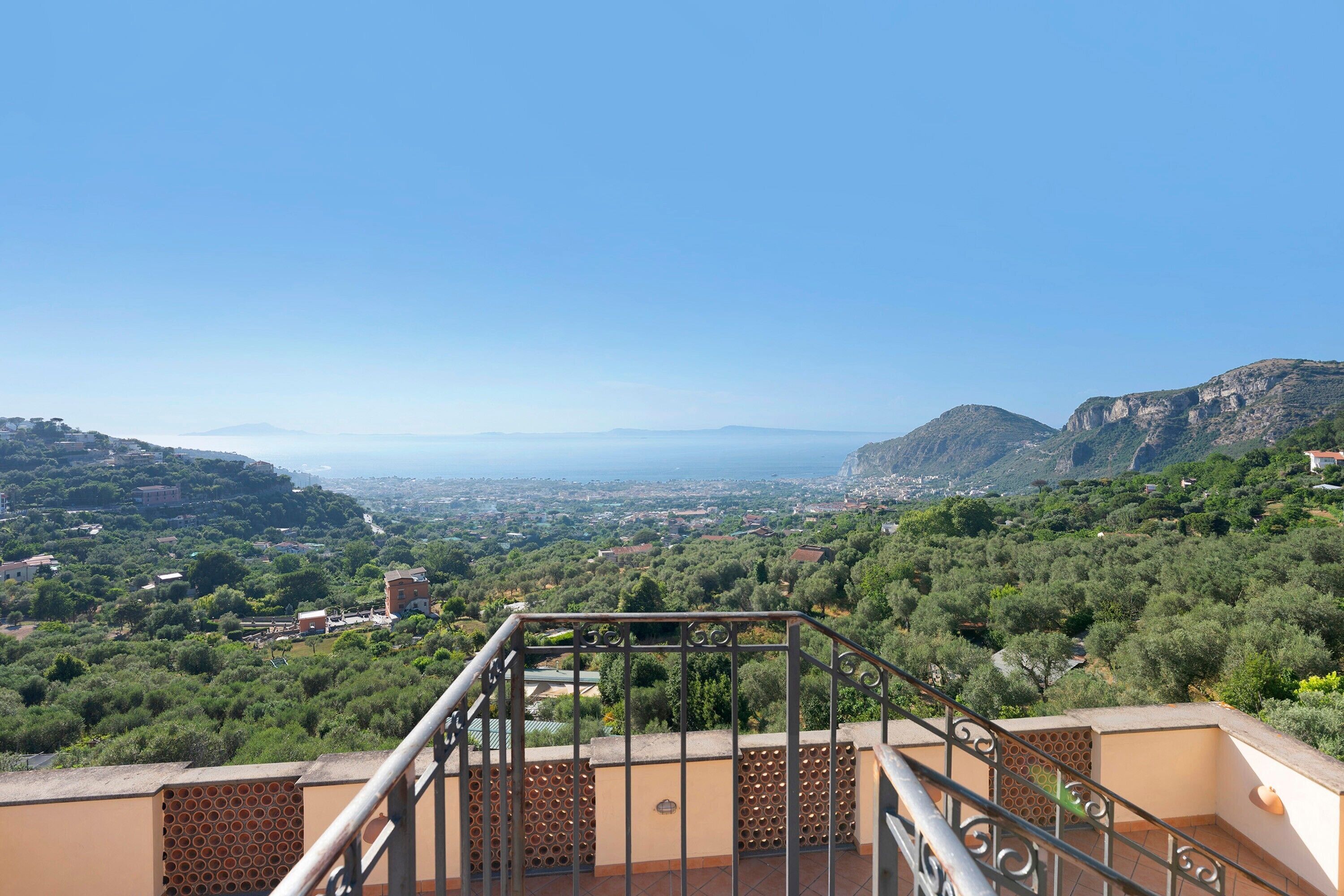 Un balcon doté d’une balustrade en métal, offrant une vue panoramique sur un paysage vallonné et, au loin, sur une ville.