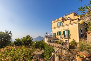 Un edificio de estilo mediterráneo con una escalera que lleva a un balcón, rodeado de vegetación exuberante y un paisaje montañoso.