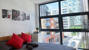 A modern bedroom with a large bed, red pillows, a nightstand, and a view of buildings through large windows. - Canary Wharf 2BR | Skyline View + Work Ready (Greater London)
