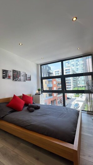 Ein modernes Schlafzimmer mit einem großen Bett, roten Kissen, einem Nachttisch und Blick auf Gebäude durch große Fenster.