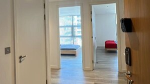 A hallway with wooden flooring, white walls, and a door with a black handle. - Canary Wharf 2BR | Skyline View + Work Ready (Greater London)