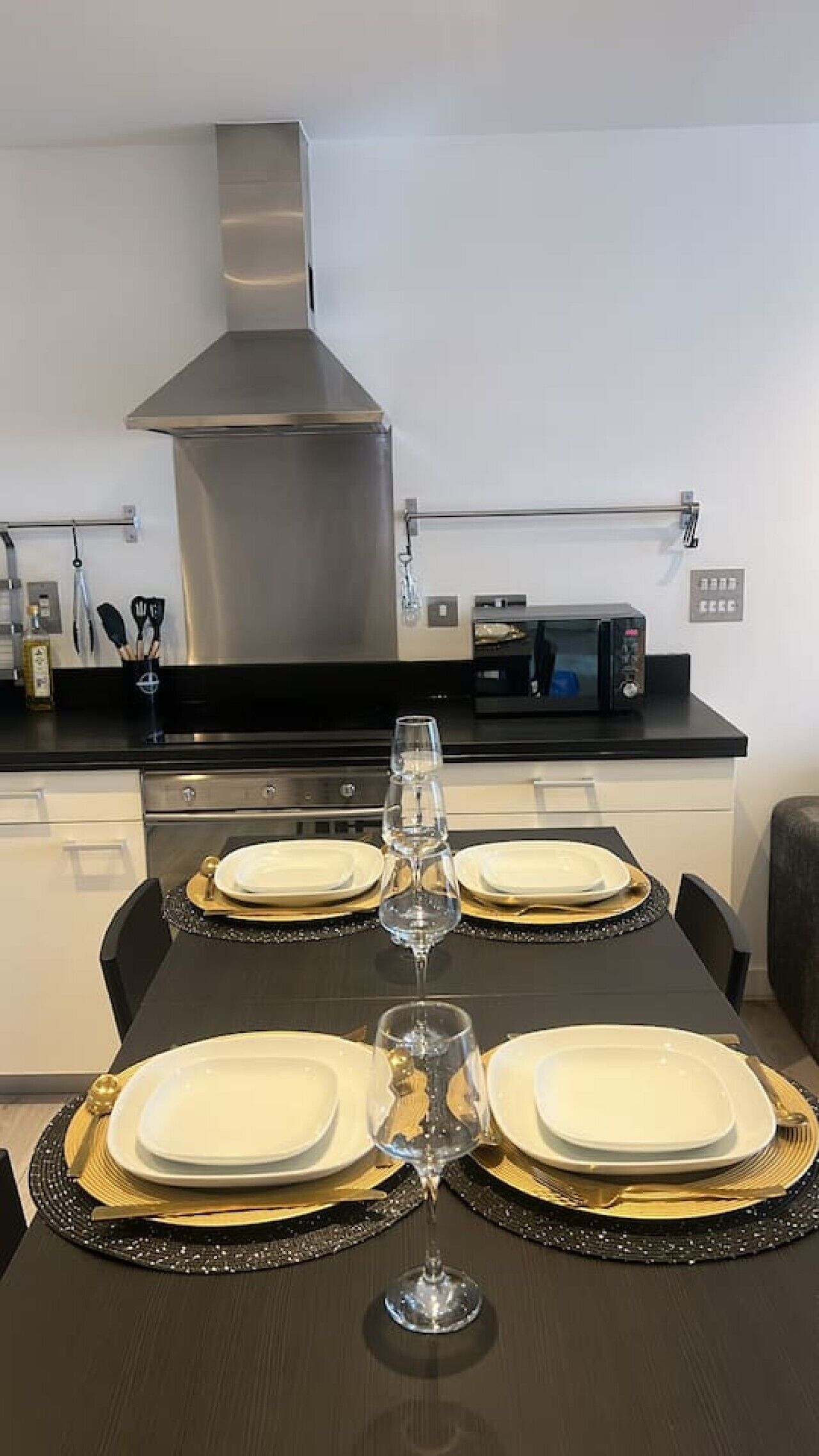 A dining table set with plates, cutlery, and wine glasses, with a microwave and cabinets in the background.