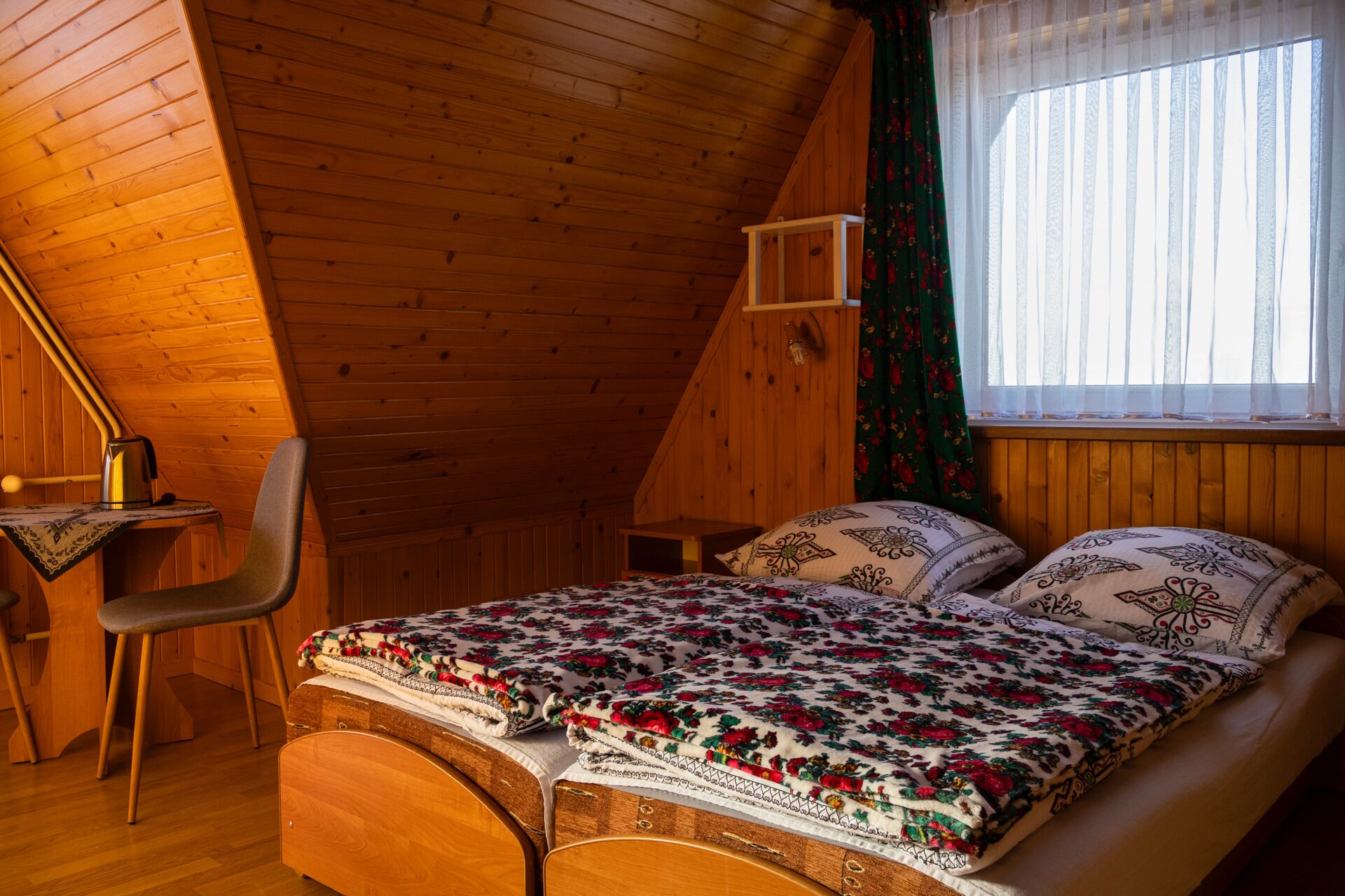 Une pièce en bois avec un plafond en pente, un lit recouvert d’une literie à motifs, une chaise et une fenêtre ornée de rideaux l égers.
