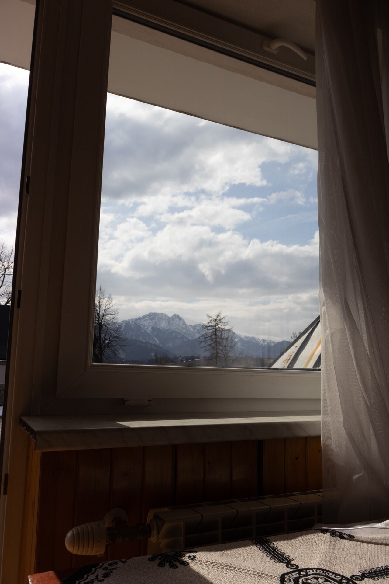 A room with a window overlooking a mountain range, a wooden bench, and a table with a patterned tablecloth.