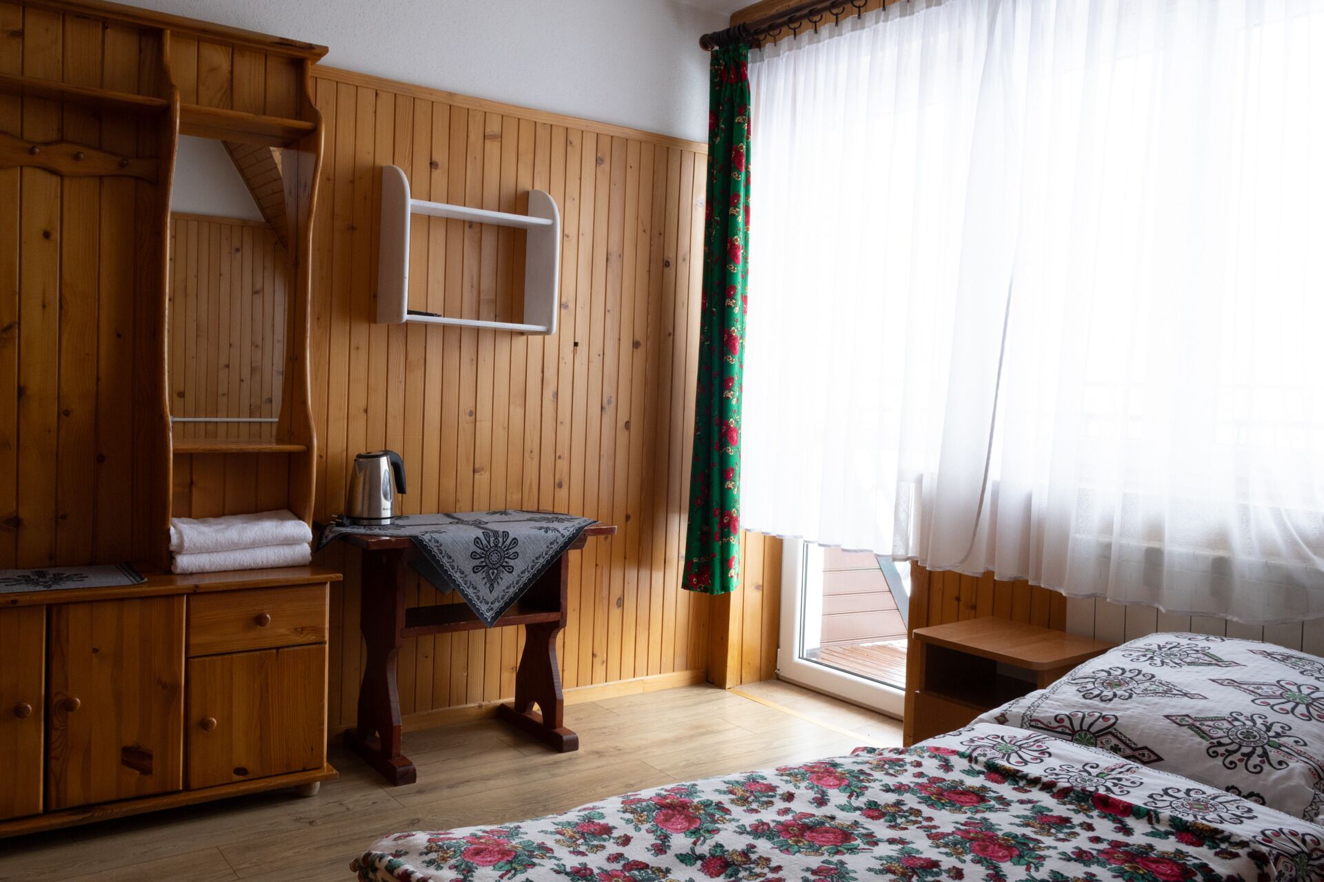 A wooden room with a bed, a table with a kettle, a shelf unit, and a window with curtains.