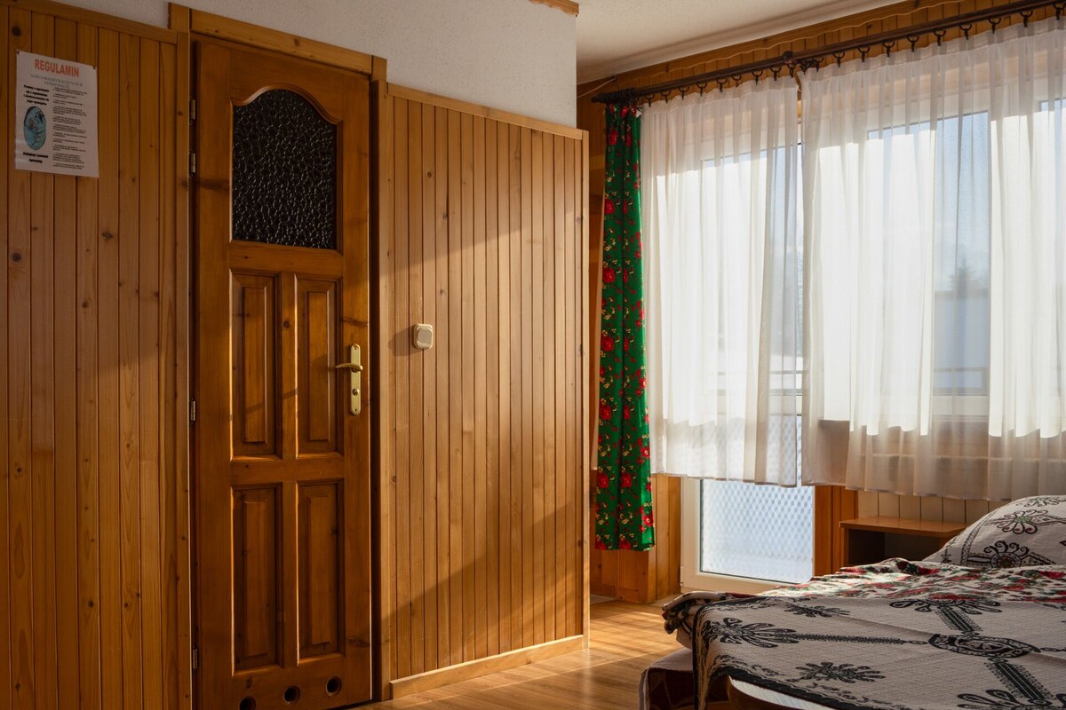 Una puerta de madera con un ventanal, una cama con cubrecama estampado y un ventanal con cortinas de gasa.
