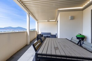 Een overdekt terras buiten, met een tafel, stoelen, een barbecue en uitzicht op verre bergen.