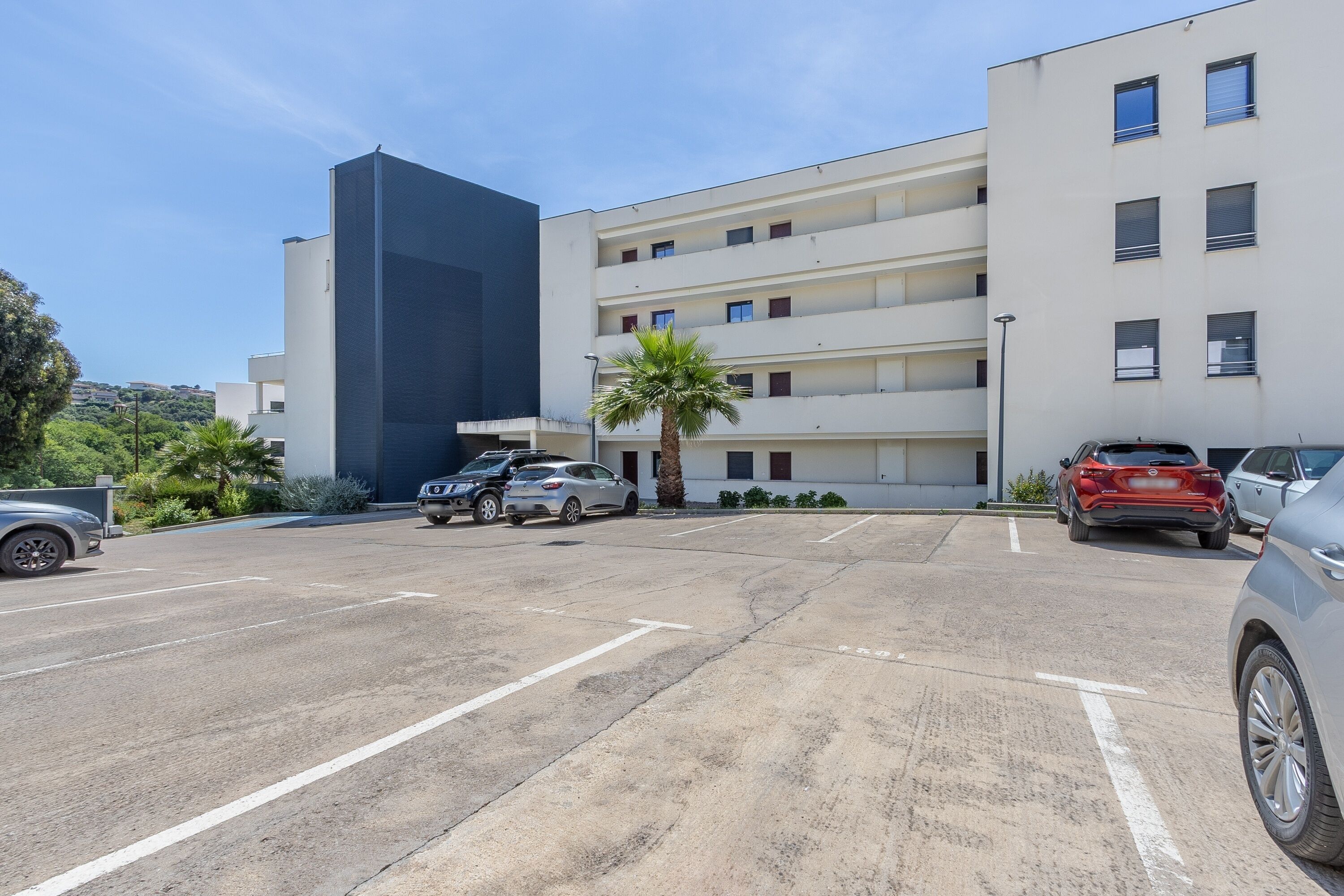 Un estacionamiento con varios coches, un edificio moderno de varios pisos y balcones, y una palmera.