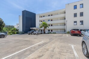 Ein Parkplatz mit mehreren Autos, ein modernes Gebäude mit mehreren Etagen und Balkonen sowie eine Palme.