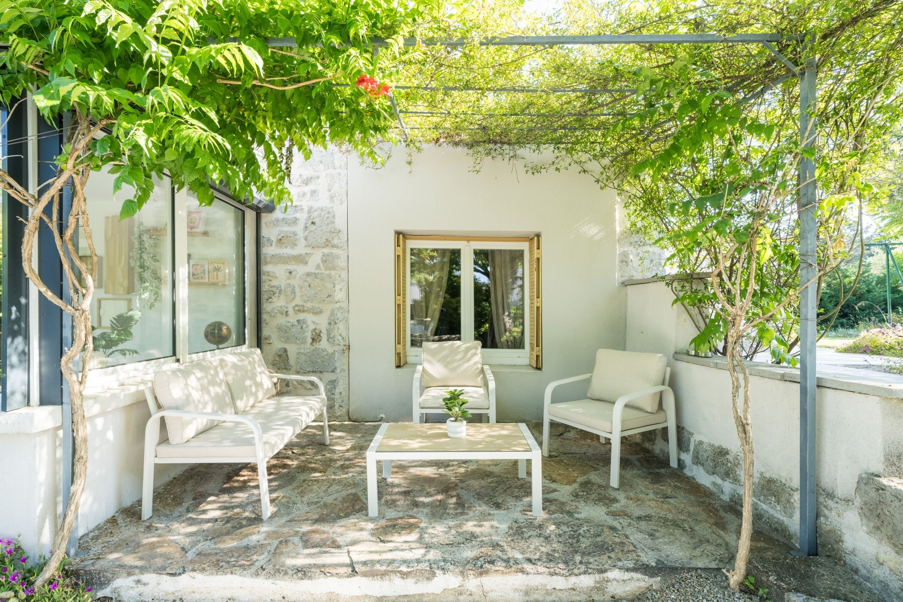 Un patio con mobili bianchi, pavimento in pietra e una pergola ricoperta di piante rampicanti.