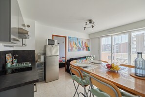 A modern kitchen with a dining area, large windows, and a colorful sailboat painting. - A l'abri côtier - Appt avec balcon proche plage (Cabourg)