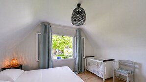 Un dormitorio con cama, cuna, ventana con cortinas, lámpara colgante y vistas a zonas verdes.
