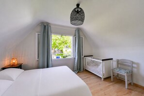 Ein Schlafzimmer mit Bett, Babybett, einem Fenster mit Vorhängen, einer Hängelampe und Blick ins Grüne.