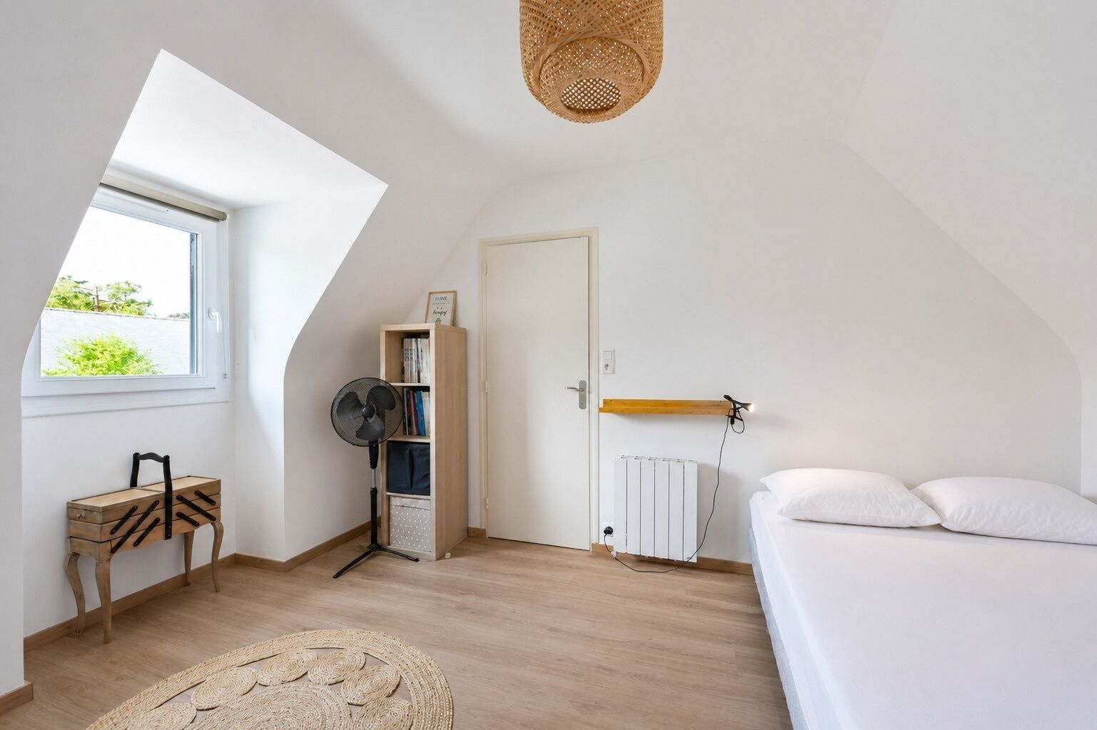 A bedroom with a sloped ceiling, a window, a radiator, a bed, a fan, a bookshelf, and a small wooden chair.