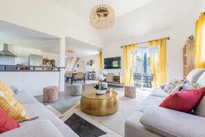 A modern living room with a sofa, ottomans, a coffee table, and a kitchen in the background. - Normandie Escape House - À 5 min des plages (Manvieux)
