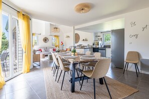 A modern kitchen with a refrigerator, stove, and dining area with a table and chairs. - Normandie Escape House - À 5 min des plages (Manvieux)