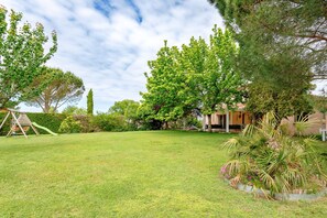 A grassy backyard with a swing set, a large tree, and a house in the background.