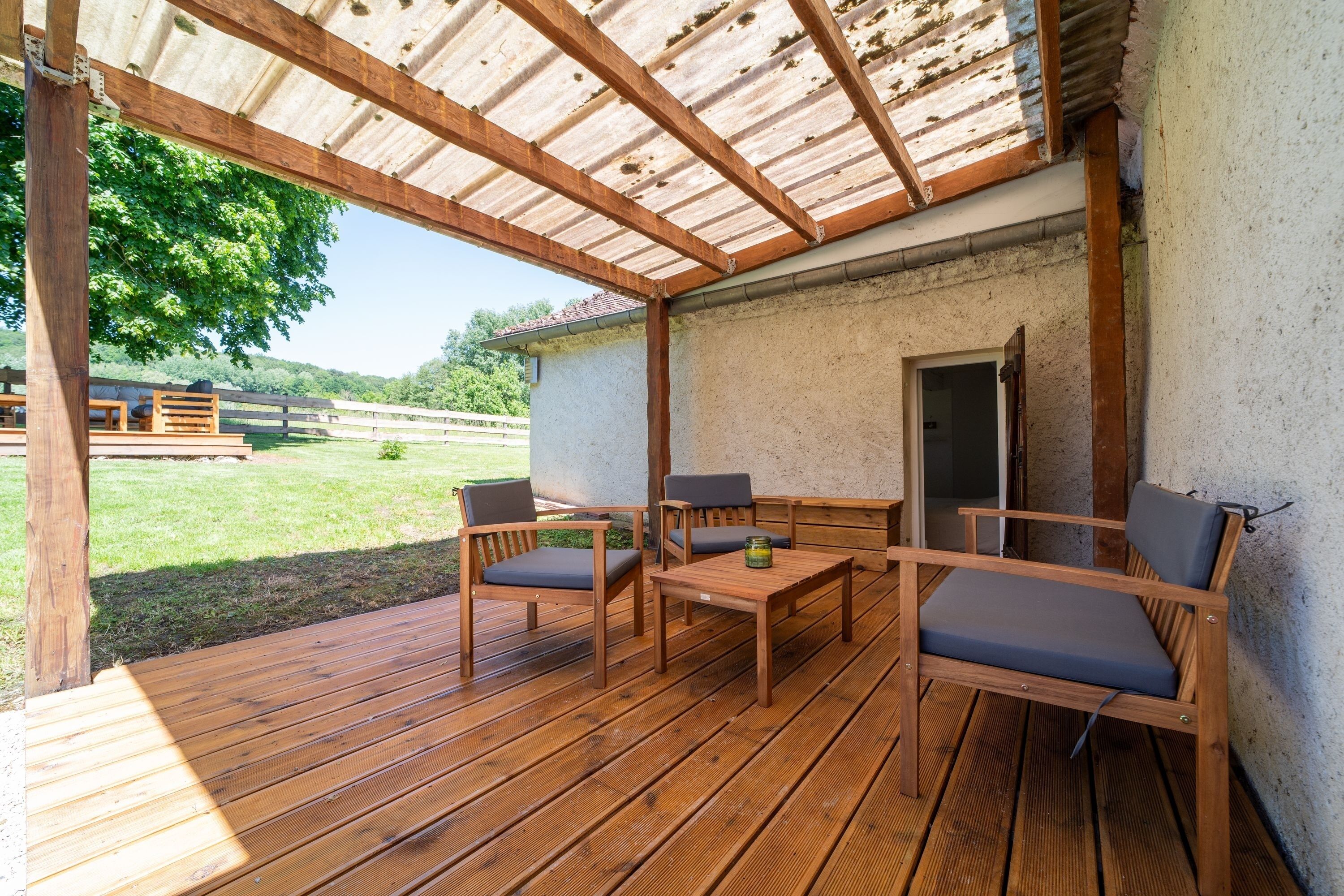 An outdoor wooden deck with a pergola, seating area, and a view of a grassy field.