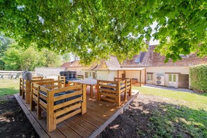 Une terrasse en bois avec des meubles d’extérieur, un bâtiment en briques en arrière-plan et un arbre qui offre de l’ombre.